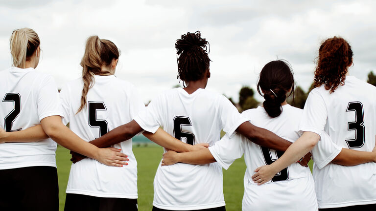 Fünf Fußballerinnen im Trikot stehen mit dem Rücken zur Kamera in einer Reihe und haben die Arme umeinander gelegt.