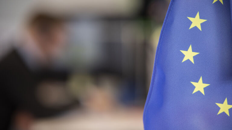 Im Vordergrund ist ein Teil der EU-Flagge zu sehen, während im Hintergrund unscharf eine Person an einem Arbeitsplatz sitzt.