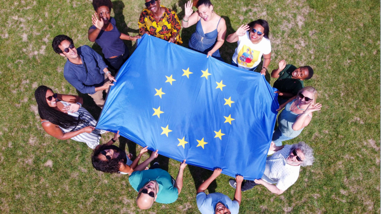 Eine Gruppe von Menschen steht um eine EU-Flagge auf einer Wiese, das Foto ist aus der Vogelperspektive aufgenommen