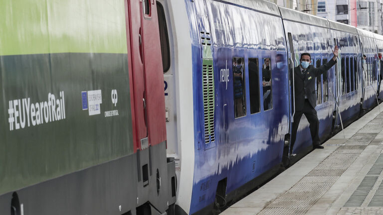 Es ist ein grün-blauer Zug zu sehen, der vom Gleis, an dem er steht, fotografiert wird. Ein Schaffner steht halb im Zug, halb auf dem Gleis.