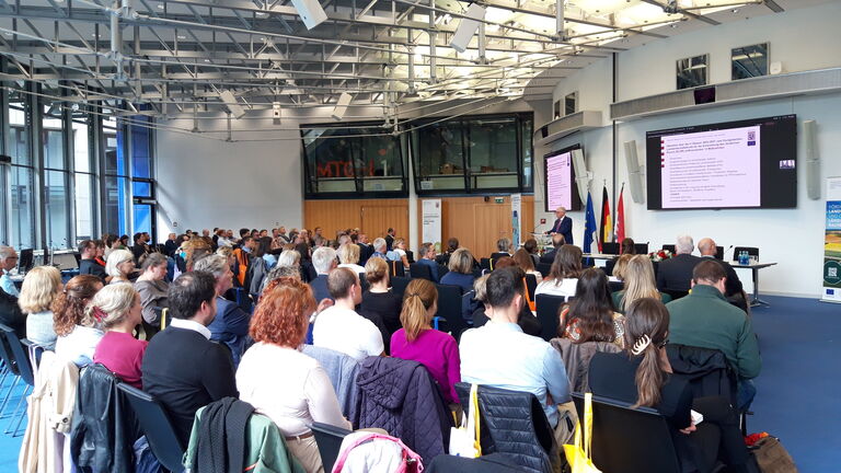 Auf dem Foto sind die Teilnehmerinnen und Teilnehmer der Förderkonferenz im Publikum von hinten zu sehen, im Hintergrund des Bildes ist ein Redner mit einer Präsentation auf einem Bildschirm zu erkennen.