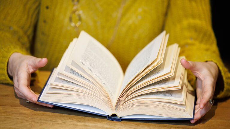 Es ist ein aufgeschlagenes Buch zu erkennen, das auf einem Holztisch liegt und von einer Frau in der Hand gehalten wird, deren Oberkörper in einem gelben Pullover zu sehen ist. 