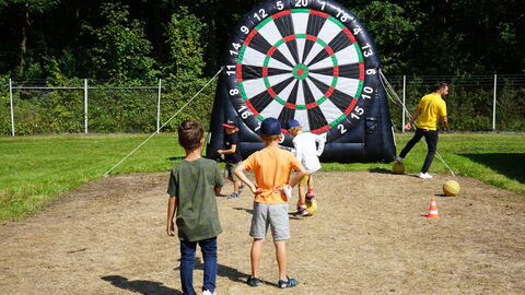 Kinder spielen auf einer Wiese im Sonnenschein vor einer großen Dartscheibe beim Drillingstreffen 2021