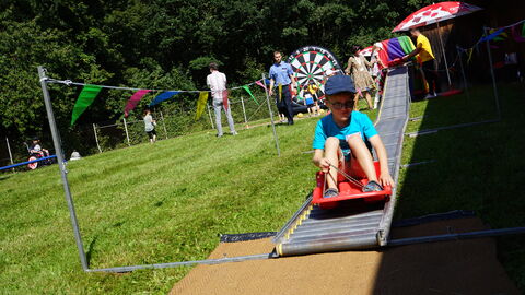 Ein Kind rodelt im Sonnenschein auf einer Wiese auf einem roten Schlitten auf einer kleinen Sommerrodelbahn beim Drillingstreffen 2021