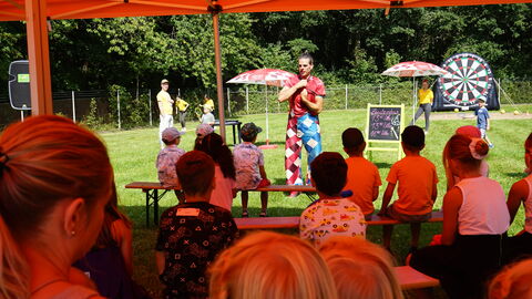 Kinder sitzen unter einen Sonnendach und schauen einer Zaubershow zu, beim Drillingstreffen 2021
