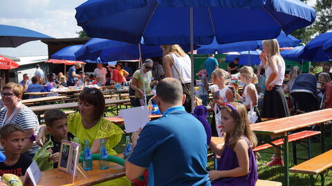 Auf Sitzbänken unter blauen Sonnenschirmen sitzen Familien beim Drillingstreffen 2021 auf einer Wiese