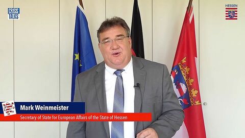 Europastaatssekretär Mark Weinmeister steht im Anzug mit Krawatte vor einem Hintergrund mit der Hessen-Fahnne und spricht