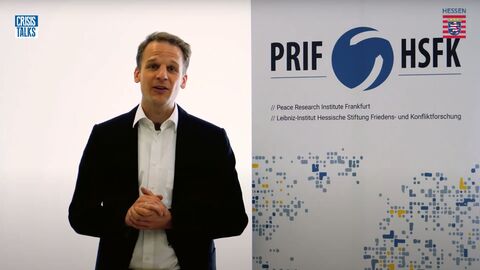 Dr. Stefan Kroll vom Leibniz-Institut Hessische Stiftung Friedens- und Konfliktforschung und Leiter Wissenschaftskommunikation steht vor einer Logo-Leinwand und spricht in die Kamera