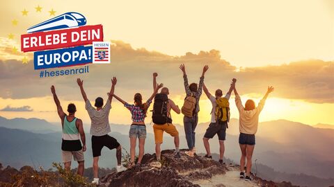 Eine Gruppe junger Menschen auf dem Gipfel eines Berges strecken die Hände gen Himmel während im Hintergrund die Sonne untergeht. 