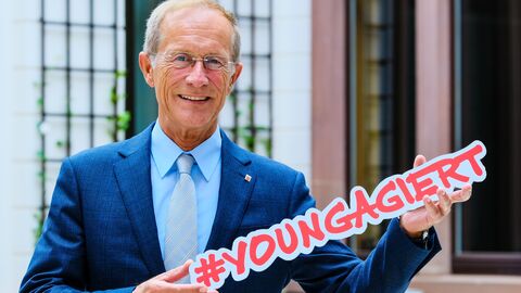 Staatsminister Wintermeyer hält den Schriftzug #youngagiert hoch und lächelt in die Kamera.