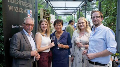 Stehen im Halbkreis mit einem Glas Wein in der Hand und lächeln in die Kamera: Mitglieder des Weinbauverbandes Hessische Bergstraße e.V. begrüßen Staatsministerin Puttrich (Mitte) und Weinkönigin Annika (2.v.r.)