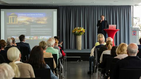 Ein Mann mit Mikrofon steht auf der Bühne vor den Zuschauern, neben ihm eine Präsentation mit Fotos der Schlösser auf einer Leinwand: Fotograf Michael Leukel verriet, wie, wann und wo seine Aufnahmen entstanden sind.
