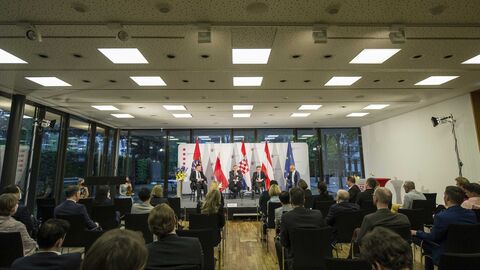 Blick in den Saal aus dem Zuschauerraum in Richtung Bühne: Man sieht die Gäste auf Stühlen sitzend auf die Bühne schauen, auf der Bühne sitzen die Teilnehmer auf Stühlen nebeneinander, hinter ihnen sind Fahnen aufgestellt (Europa, Hessen, weitere Länder-Fahnen)