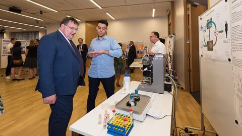 Ein Mann und ein Jugendlicher stehen vor einem Tisch mit Laborgegenständen: Christos Assiklaris (17) aus Offenbach präsentiert Europastaatssekretär Mark Weinmeister stolz sein Projekt "Insektenpathogene Pilze und ihr Potenzial Kunststoff biologisch abzubauen“