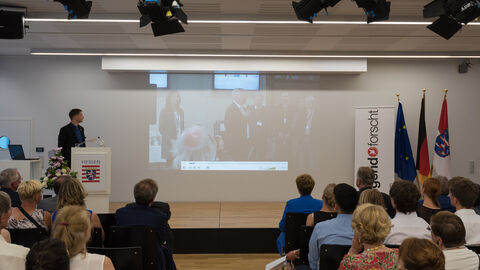 Ein Mann steht auf einer Bühne, hinter ihm ist eine Präsentation zu sehen. Im Vordergrund sieht man einen Teil des Publikums von hinten: Oliver Karplak, Landeswettbewerbsleiter Jugend forscht Hessen