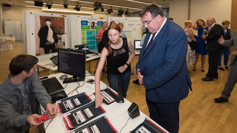 Ein Mann und eine Jugendliche stehen vor einem Tisch mit Heften und einem Computer darauf: Ferdinand Krämer (21) und Klara Krämer (18) aus Weitersburg haben einen eigenen Sprachassistenten konstruiert „TIANE – ein offener Smart-Home-Sprachassistent“, den sie Europastaatssekretär Mark Weinmeister bei seinem Rundgang vorstellen