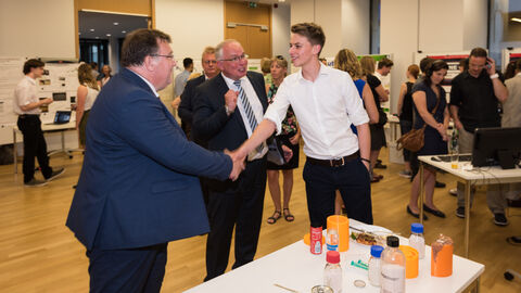 Ein Mann und ein Jugendlicher schütteln sich die Hand. Vor ihnen steht ein Tisch mit Gegenständen darauf: 