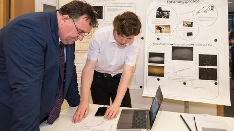 Ein Man und ein Jugendlicher sind über einen Laptop gebeugt, der auf einem Tisch steht: Europastaatssekretär Mark Weinmeister erörtert mit den Jungforscherinnen und Jungforschern die Forschungsergebnisse, hier mit Moritz von Eimern (18) über die Vermessung der LBV Sterne Deneb und P Cyghni