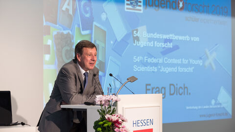 Ein Mann in einem Anzug steht auf einer Bühne hinter einem Podium. Im Hintergrund ist eine Präsentation zu sehen: Jürgen Tiedje, Europäische Kommission, Generaldirektion Forschung und Innovation