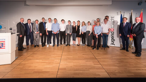 Junge Erwachsene und Erwachsene stehen nebeneinander auf einer Bühne und lächeln in die Kamera. Rechts neben ihnen steht ein Banner mit der Aufschrift "jugend forscht"