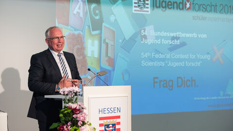 Ein Mann in einem Anzug steht auf einer Bühne hinter einem Podium. Im Hintergrund ist eine Präsentation zu sehen: Dr. Sven Baszio, Geschäftsführer Vorstand Stiftung Jugend forscht e.V