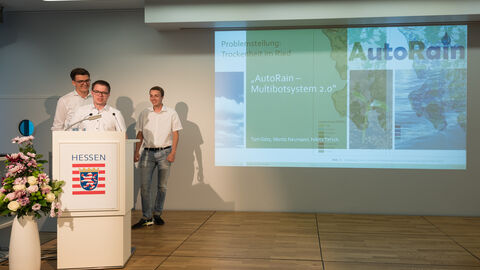 Auf einer Bühne stehen drei Jugendliche, die alle ein weißes Hemd tragen. Hinter ihnen ist eine Präsentation zu sehen: Projekt "AutoRain": Als Vertreter des Hessischen Landeswettbewerbs haben (v.l.n.r.) Tom Götz (18), Moritz Neumann (18), Nikita Tatsch (19) ihr Projekt „AutoRain 2.0“ präsentiert. Die Schüler des Lessing-Gymnasium Lampertheim haben ein Wechselträgersystem entwickelt, womit Landwirte bei vielseitigen Arbeitsgängen, wie z.B. Düngerausbringung, in ihrem Alltag entlastet werden können
