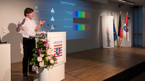 Ein Jugendlicher steht auf einer Bühne und hält eine Präsentation: Moritz von Eimern (18) aus Wuppertal erklärt die „Spektroskopische Vermessung der LBV-Sterne Deneb und P Cygni“, um die Geschwindigkeit der Sternwinde zu ermitteln