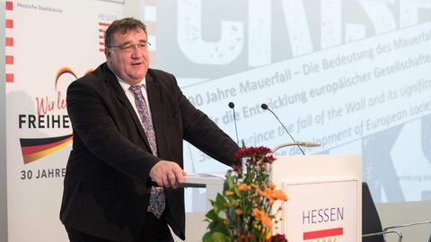 30 Jahre Mauerfall: Die Bedeutung des Mauerfalls für die Entwicklung europäischer Gesellschaften Ein Mann im Anzug steht auf einer Bühne hinter einem Podium. Hinter ihm ist eine Präsentation zu sehen.
