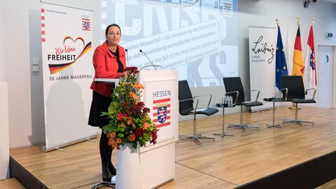 Eine Frau steht auf einer Bühne an einem Podium mit dem Hessenwappen darauf. Hinter ihr ist ein Banner mit der Aufschrift "Wir leben Freiheit - 30 Jahre Mauerfall" und eine Präsentation zu sehen