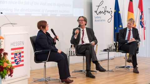 30 Jahre Mauerfall: Die Bedeutung des Mauerfalls für die Entwicklung europäischer Gesellschaften Zwei Männer und eine Frau sitzen auf Stühlen auf einer Bühne. Sie haben ein Mikrofon in der Hand und hinter ihnen ist eine Präsentation zu sehen