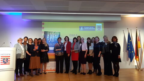 Empfang der European Women Lawyers Association Frauen stehen auf einer Bühne mit einer Präsentation im Hintergrund und lächeln in die Kamera:
