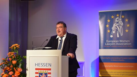 Empfang der European Women Lawyers Association Ein Mann in einem Anzug steht auf einer Bühne hinter einem Podium mit dem Hessenwappen darauf. Hinter ihm ist ein Banner der  European Women Lawyers Association zu sehen