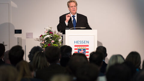 Ein Mann in einem Anzug steht vor einem Podium auf einer Bühne. Im Vordergrund sind die Hinterköpfe von Menschen aus dem Publikum zu sehen: Ansprache Martin Kotthaus, Botschafter der Bundesrepublik Deutschland beim Königreich Belgien