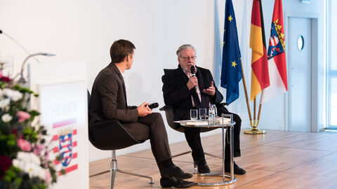 Zwei Männer in Anzügen sitzen auf einer Bühne und haben Mikrofone in der Hand. Im Hintergrund sind die Hessen-, Deutschland- und Europaflagge zu erkennen: Gunnar Krüger, ZDF Korrespondent, Studio Brüssel, im Gespräch mit dem Zeitzeugen, dem damaligen Klassensprecher, Karsten Köhler
