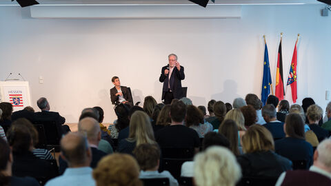 Zwei Männer in Anzügen sind auf einer Bühne und haben Mikrofone in der Hand. Im Vordergrund sind Hinterköpfe des Publikums zu sehen: Karsten Köhler beantwortet Fragen der Teilnehmer 