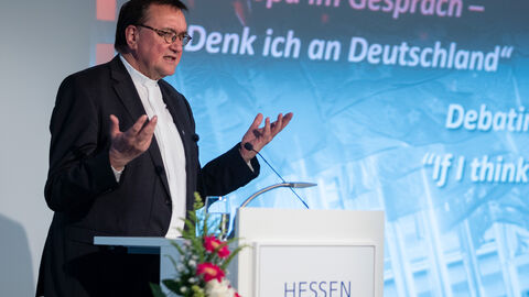 Ein Mann in einem Anzug steht hinter einem Podium. Im Hintergrund ist eine Präsentation zu sehen: Prof. Dr. Martin Hein, Bischof der Evangelischen Kirche von Kurhessen-Waldeck