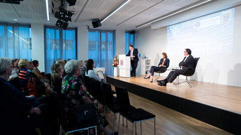 Von der Seite sieht man drei Personen auf einer Bühne. Zwei davon sitzen und einer steht hinter einem Podium, links im Bild sieht man Teile des Publikums: v.l.n.r.: Mark Weinmeister, Hessischer Staatssekretär für Europa, Franziska Broich und Prof. Dr. Martin Hein