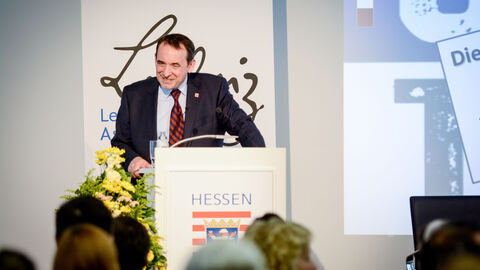 Ein Mann in einem Anzug steht hinter einem Podium: Der Hessische Kultusminister Prof. Dr. R. Alexander Lorz begrüßte im Namen der Hessischen Landesregierung die Gäste