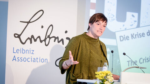 Eine Frau mit einem grünen Oberteil steht hinter einem Podium: Begrüßung durch Prof. Dr. Nicole Deitelhoff, Direktorin des Leibniz-Instituts Hessische Stiftung Friedens- und Konfliktforschung/Sprecherin des Leibniz-Forschungsverbunds „Krisen einer globalisierten Welt"