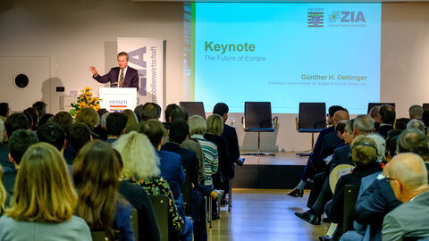 Es sind Zuschauer von hinten zu sehen, die einen Mann auf einer Bühne anschauen. Der Mann trägt einen dunklen Anzug und steht hinter einem Podium mit dem Hessenwappen drauf. Rechts daneben ist eine Präsentation zu sehen mit dem Titel "Keynote - The Future of Europe".