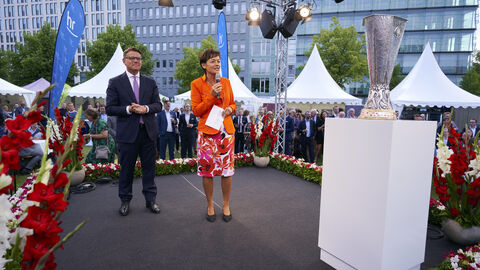 Der Hessenabend beginnt. Ministerpräsident Boris Rhein und Staatsministerin Lucia Puttrich begrüßen die Gäste: Boris Rhein (links) und Lucia Putrich (rechts, mit Mikrofon) stehen auf einer mit Blumen geschmücken Bühne und sprechen zu den Gästen