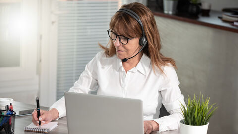 Eine Frau mit Headset sitzt vor einem Laptop und schreibt auf einem Notizblock.