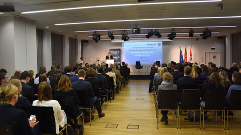 Im Vordergrund sind Personen von hinten zu sehen, die in Stuhlreihen sitzen und auf eine Bühne schauen. Im hinteren Teil des Bildes sieht man eine Bühne auf der ein Mann im Anzug hinter einem Podium steht. Hinter ihm sieht man eine Präsentation mit dem Titel "EU-Förderperiode nach 2020 – Herausforderungen und Erfordernisse"