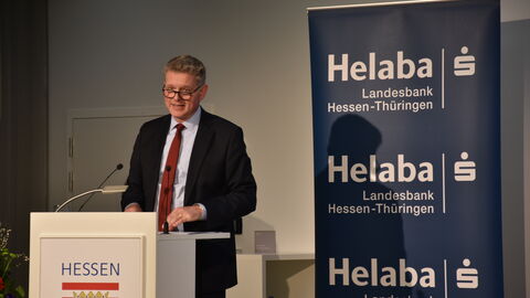 Ein Mann in einem Anzug steht auf einer Bühne hinter einem Podium auf dem das Hessenwappen zu sehen ist. Neben ihm ist ein Banner aufgestellt mit dem Schriftzug "Helaba Landesbank Hessen-Thüringen" und dem Logo der Sparkasse.