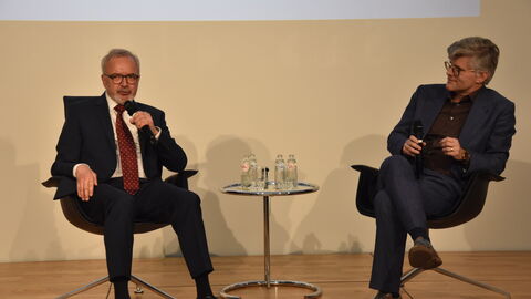 Zwei Männer in dunkeln Anzügen sitzen auf Stühlen nebeneinander. Zwischen ihnen befindet sich ein Tisch mit Wasserflaschen. Beide Männer halten ein Mikrofon in der Hand und hinter ihnen ist eine Präsentation zu sehen.