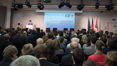 Begrüßung von Staatsministerin Lucia Puttrich Auf einer Bühne steht eine Frau, die ein blaues Oberteil trägt, hinter einem Podium mit dem Hessenwappen drauf. Eine Präsentation mit dem Titel "Welcome: Lucia Puttrich" ist rechts von ihr zu sehen. Im Vordergrund sieht man die Hinterköpfe der Zuschauer, die Richtung Bühne schauen.