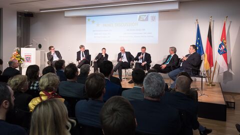 Auf einer Bühne sitzen sieben Männer in dunklen Anzügen auf Stühlen nebeneinander. Einige von ihnen haben Mikrofone in der Hand. Links im Bild steht ein Podium mit dem Hessenwappen drauf und rechts im Bild sieht man die Europa-, Deutschland- und Hessenflagge. Im Vordergrund sieht man die Hinterköpfe der Zuschauer.