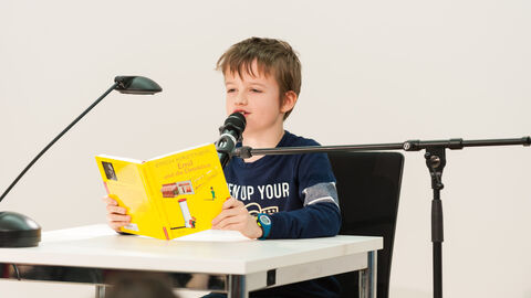 Vorlesewettbewerb 2019 Ein Junge mit hellbraunen Haaren und einem blauen Shirt sitze an einem Tisch mit einem Buch in der Hand. Er spricht in ein Mikrofon.