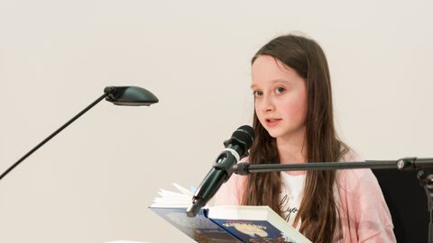 Vorlesewettbewerb 2019 Ein Mädchen mit braunen Haaren und einem rosa Oberteil sitzt an einem Tisch mit einem Buch in der Hand. Sie spricht in ein Mikrofon.