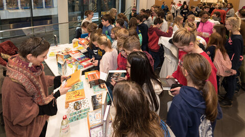 Vorlesewettbewerb 2019 Eine Frau sitzt mit einem Buch in der Hand an einem Tisch auf dem viele weiter Bücher liegen. Davor stehen Kinder, die ich die Bücher anschauen.